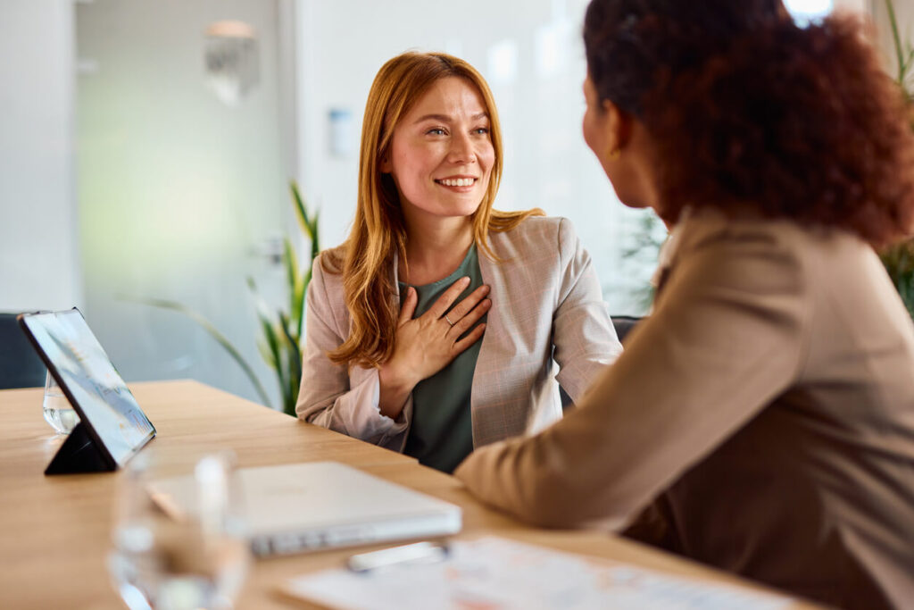 Two smiling businesswomen sitting at a modern office table, engaging in a friendly conversation.